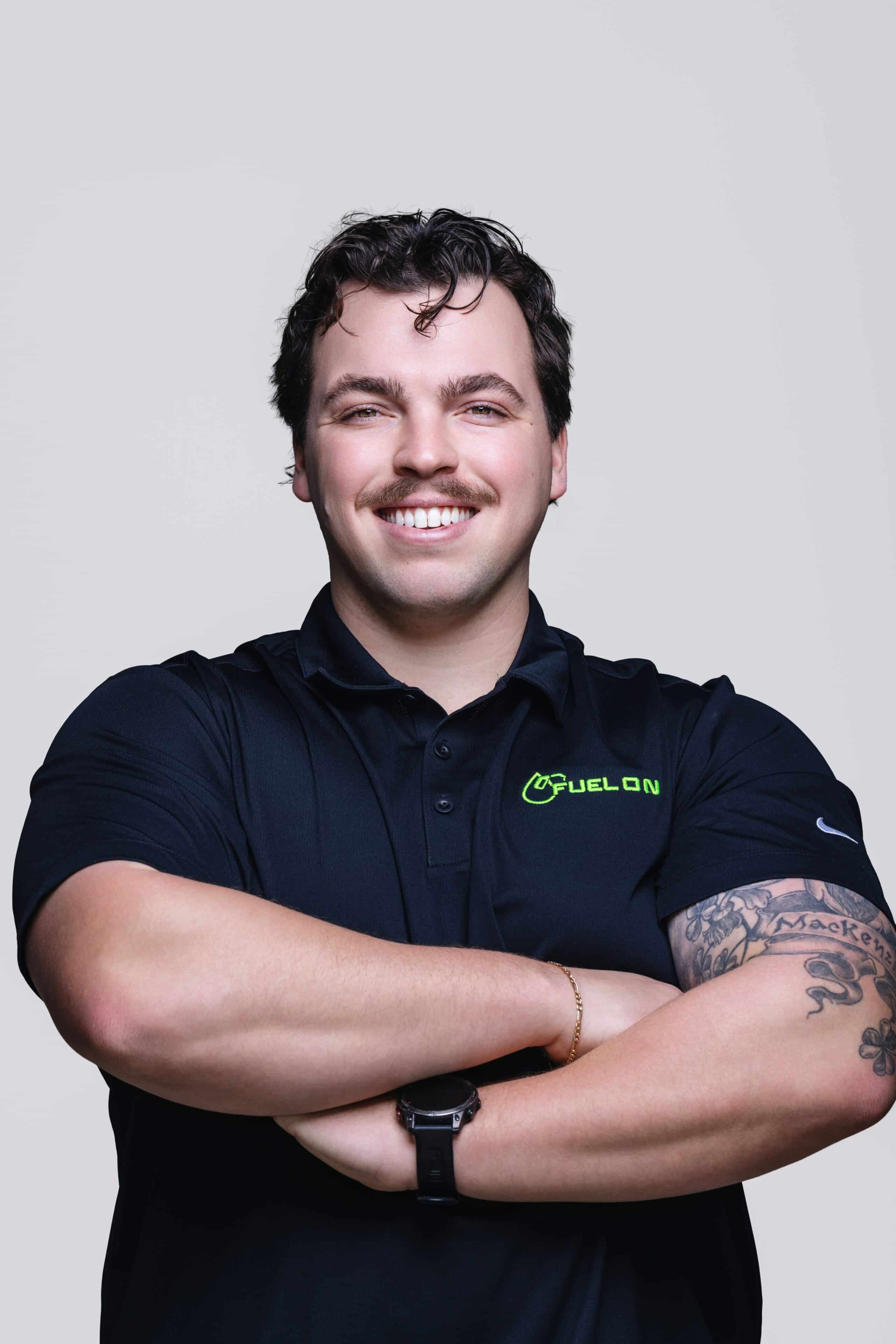 Smiling man with dark hair and mustache wearing a black polo shirt with a green FUEL ON logo, arms crossed, showing tattoos on his right forearm against a light background.