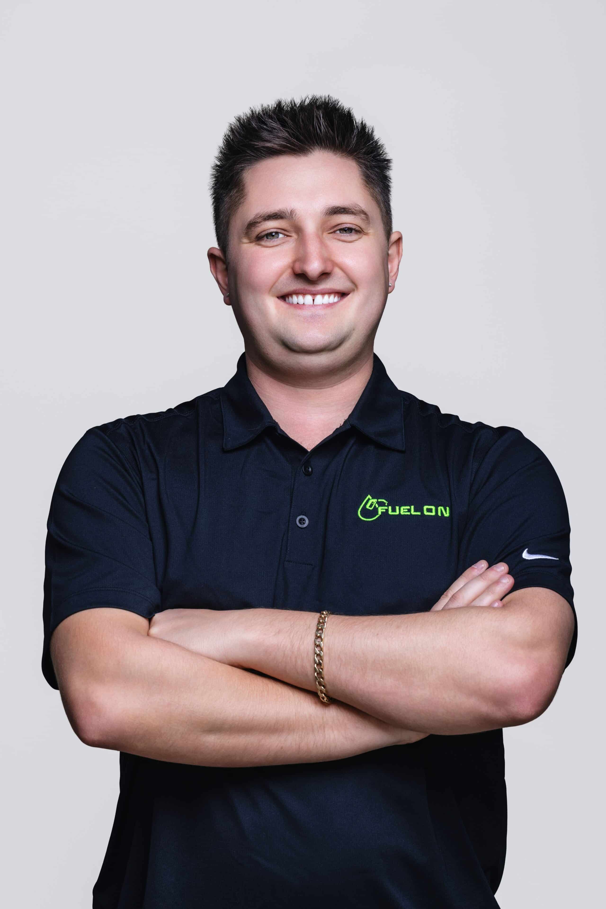 A man with short brown hair stands smiling with arms crossed, wearing a black collared shirt with a FuelOn logo and a gold bracelet against a plain light background.