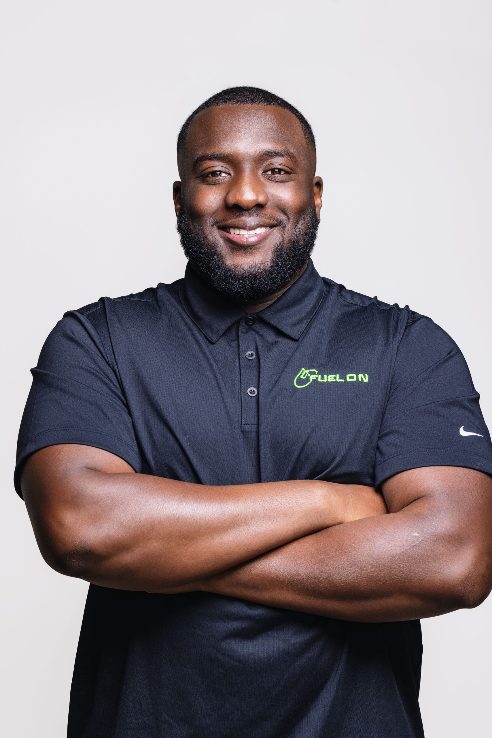 Man wearing a black polo shirt with FUEL ON logo, standing with arms crossed and smiling in front of a plain light background.