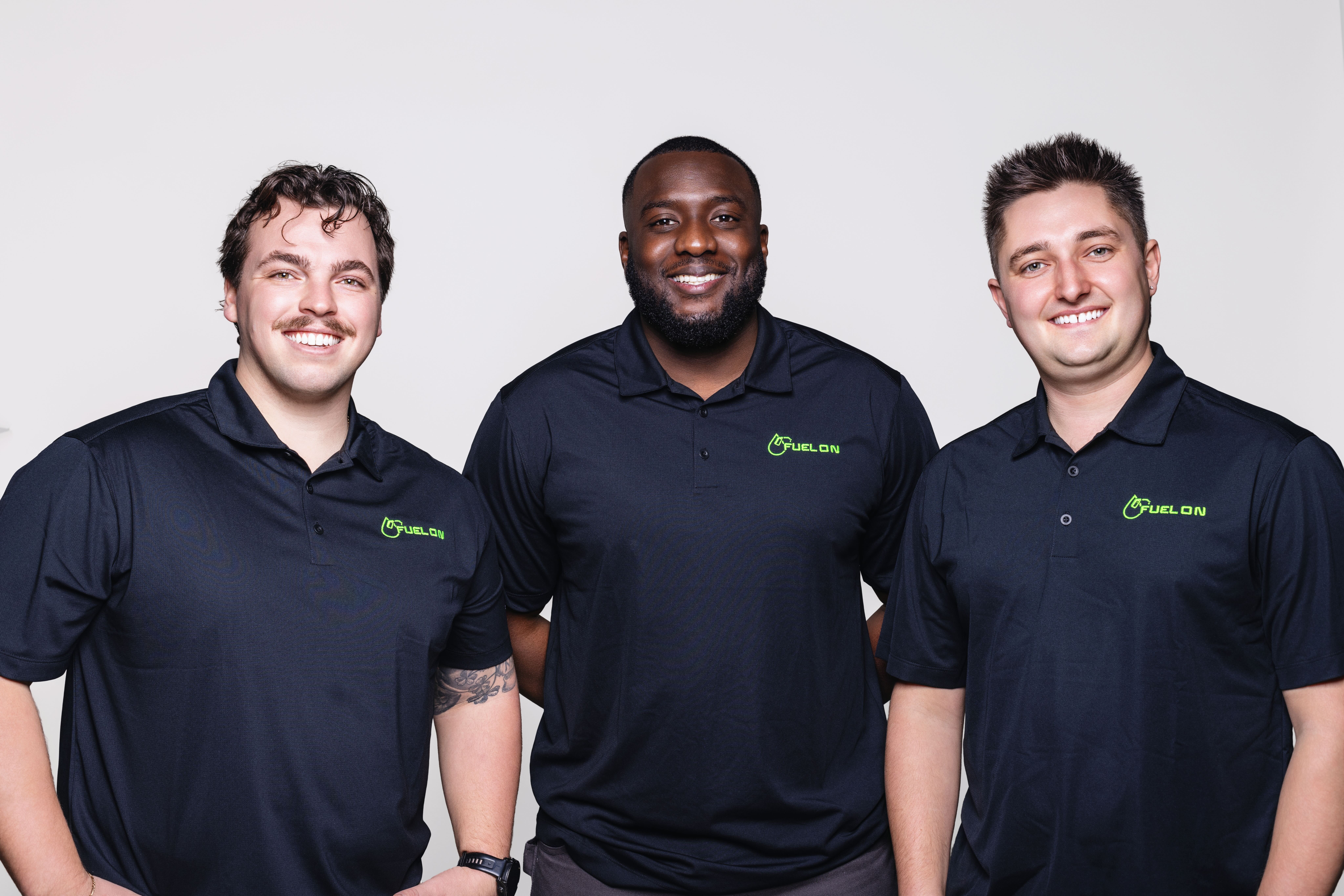 Three men stand side by side, smiling at the camera, wearing matching black polo shirts with a green FuelOn logo on the chest.